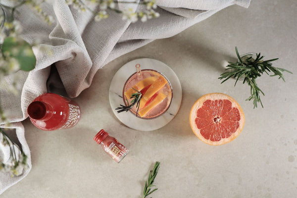 Una limonata al pompelmo e melograno in un bicchiere, decorato con un pompelmo a fette, rametti di timo e il Ginger Shot Pomegranate di Kloster Kitchen.