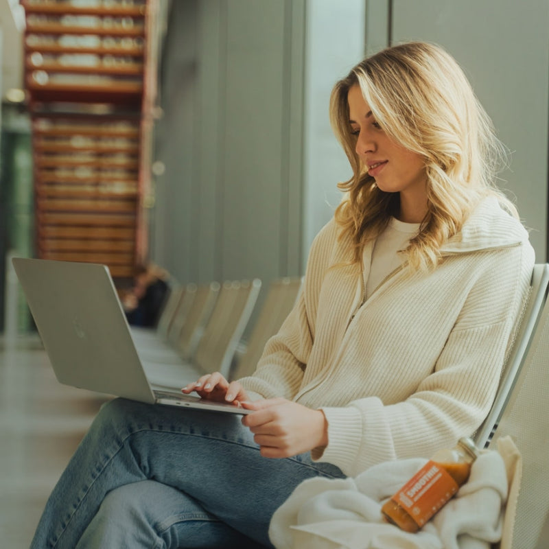 Une femme est assise sur une chaise, son ordinateur portable ouvert sur ses genoux. Un smoothie Kloster Kitchen est posé à côté d'elle.